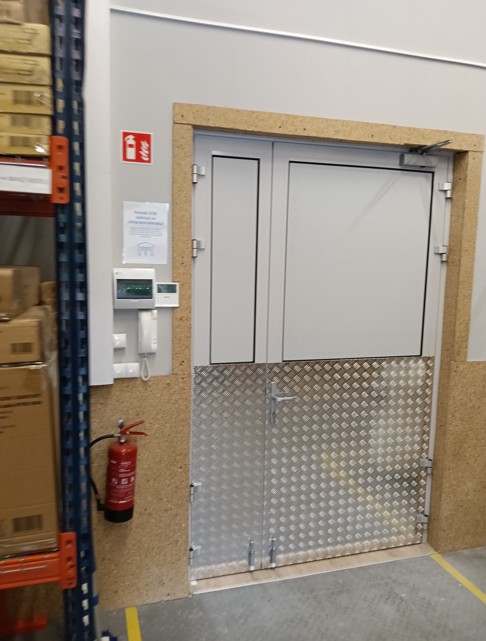 Szare, dwuskrzydłowe drzwi techniczne z aluminiowym zabezpieczeniem dolnej części, zamontowane w magazynie. Obok gaśnica i system alarmowy.