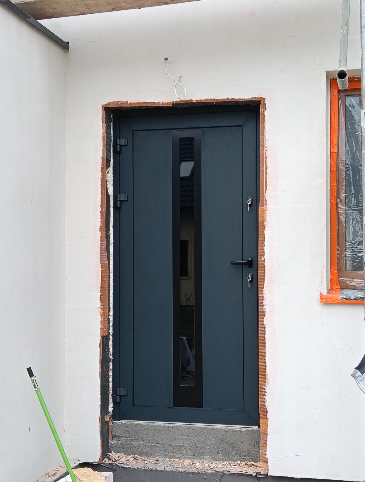 Montaż grafitowych drzwi zewnętrznych z szybą w surowym otworze budynku. Widoczne zabezpieczone okno i narzędzie. Etap prac wykończeniowych.