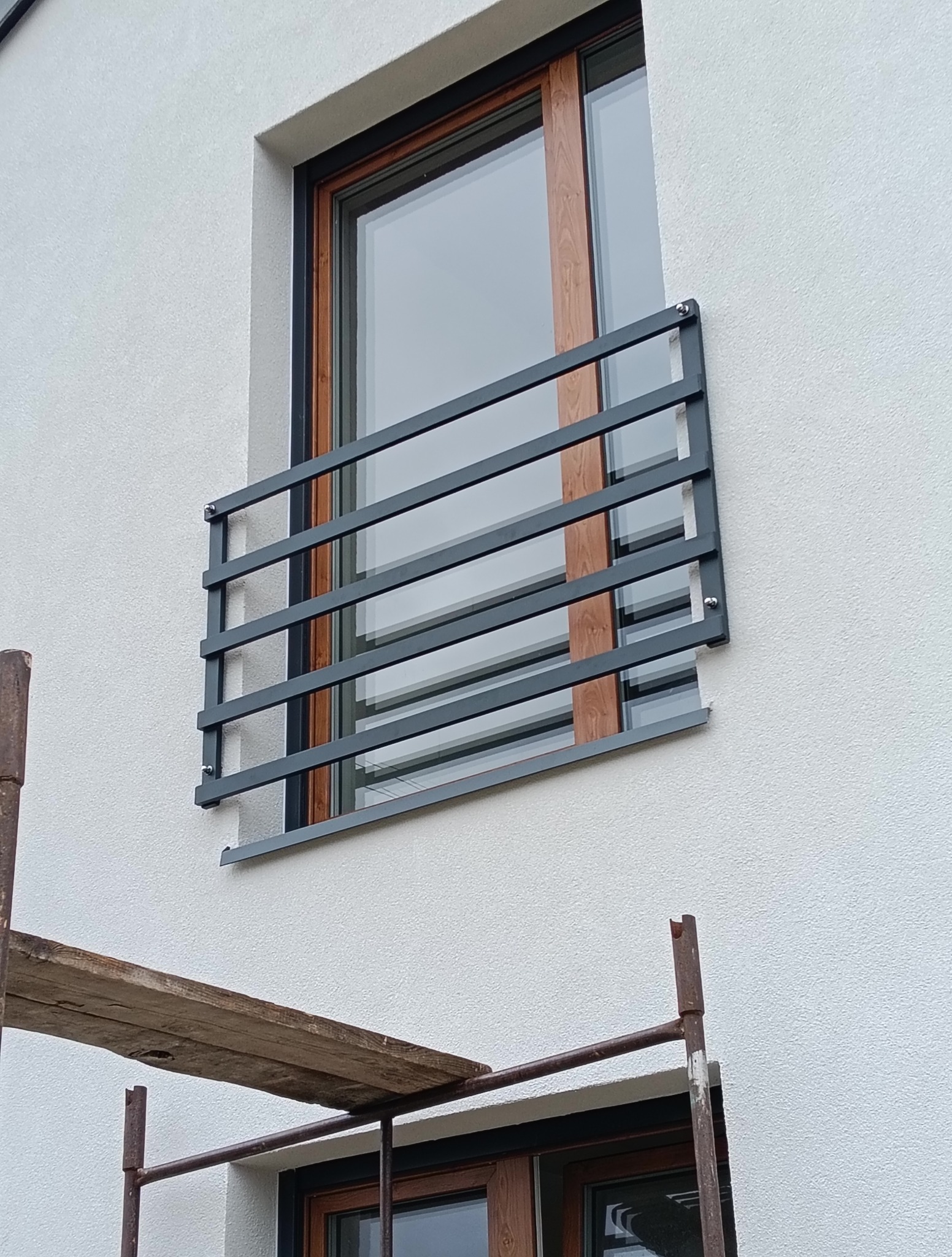 Okna z drewnopodobną ramą i szarymi, poziomymi balustradami zamontowanymi na elewacji budynku. Fragment rusztowania w kadrze.