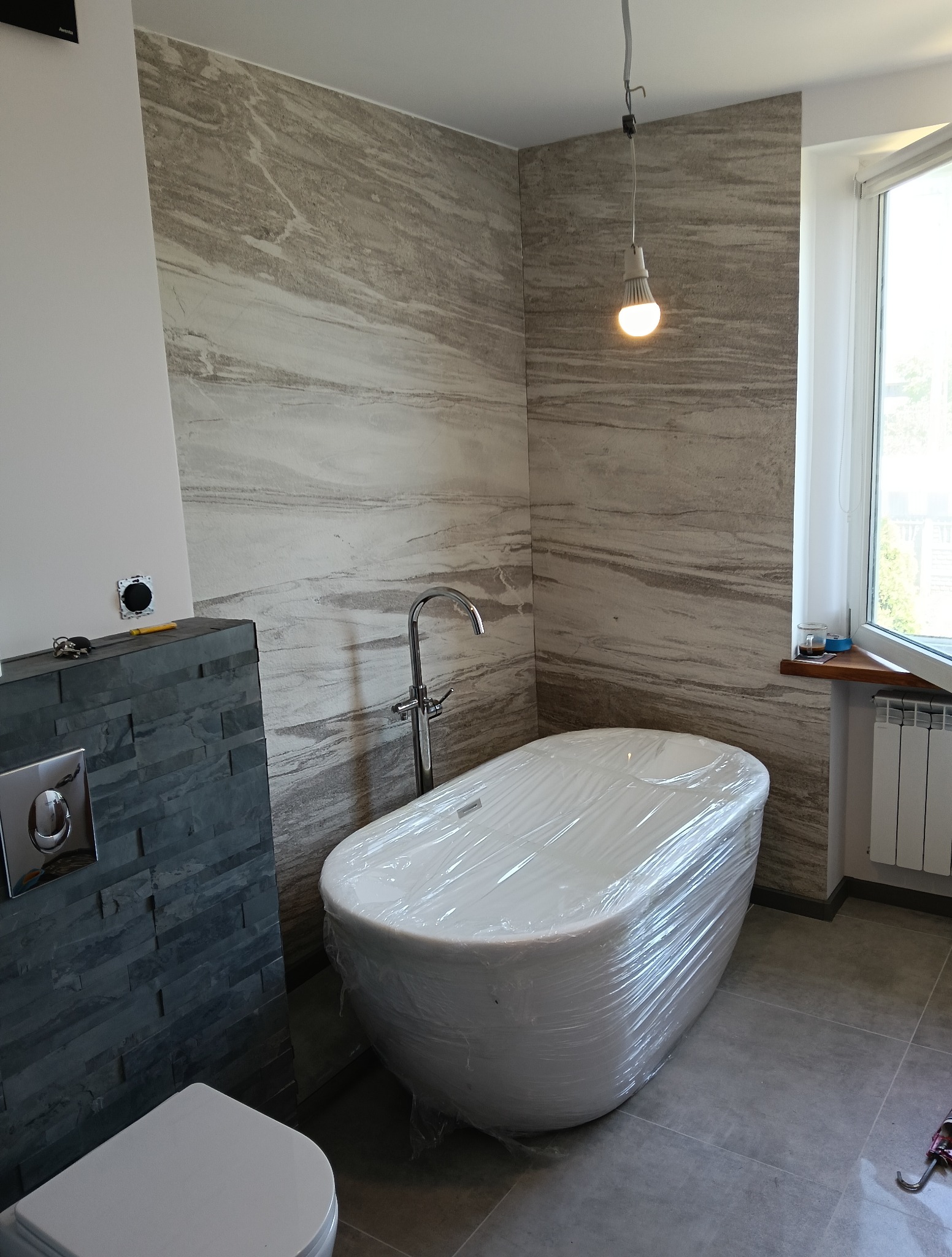 Nowoczesna łazienka w trakcie wykończenia: wanna owinięta folią ochronną, szara ściana z kamienia, bateria stojąca i okno z grzejnikiem. Widoczna surowa żarówka.