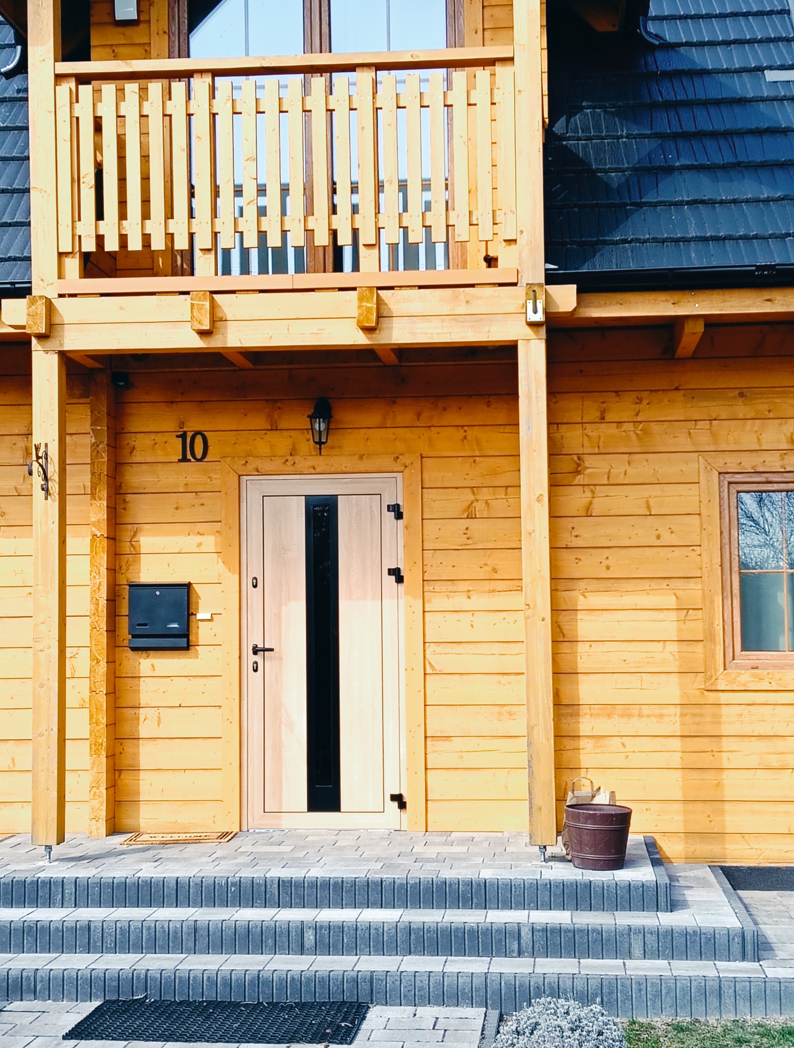 Drewniany dom z balkonem, jasnymi drzwiami z czarnym paskiem i numerem 10. Widoczne schody z kostki brukowej i metalowa skrzynka na listy.