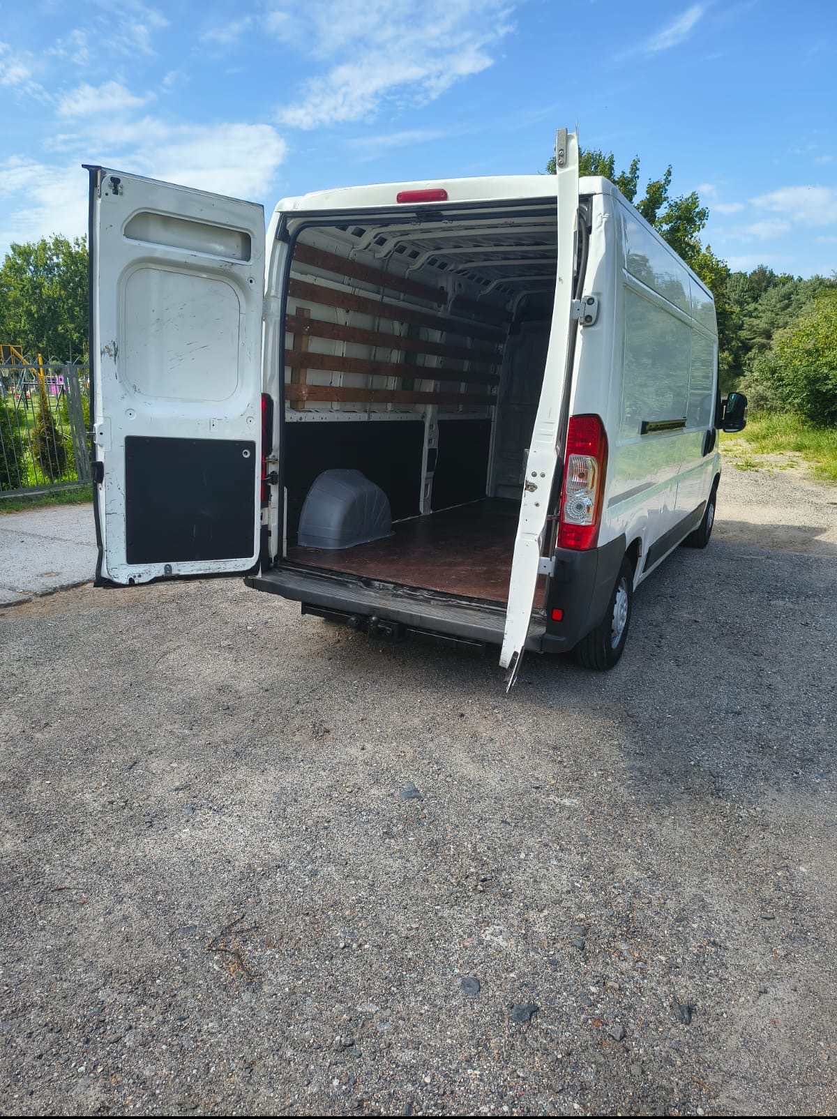 Biały bus z otwartymi tylnymi drzwiami, pusta przestrzeń ładunkowa z drewnianymi listwami, widok z tyłu na żwirowym podłożu i zieleń w tle.