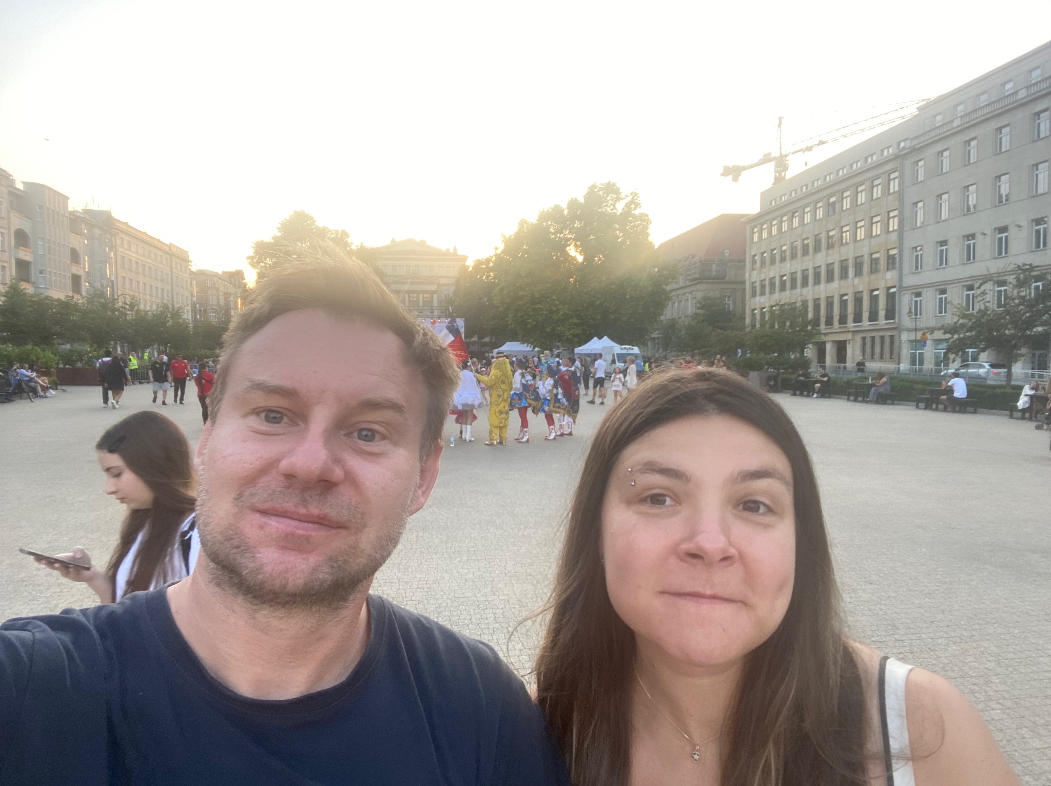 Para robi sobie selfie na tle miejskiego placu z budynkami w tle. Widoczny tłum ludzi i budowa z żurawiem. Słoneczny dzień.