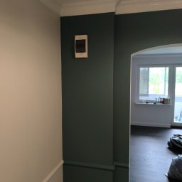 Łukasz Oczeretko Usługi Ogólnobudowlane - Ściana pomalowana na dwa kolory z dekoracyjną listwą i skrzynką elektryczną. Widok na pokój z oknem i podłogą z paneli. Częściowo widoczny odkurzacz.