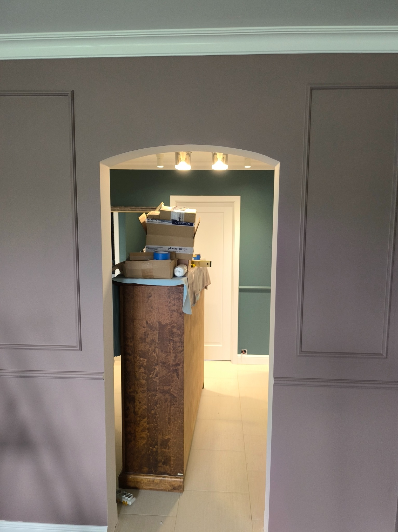 Wykończone wnętrze z szarą ścianą z dekoracyjnymi panelami, widok przez łuk na zieloną ścianę i białe drzwi. Na pierwszym planie komoda z kartonami.