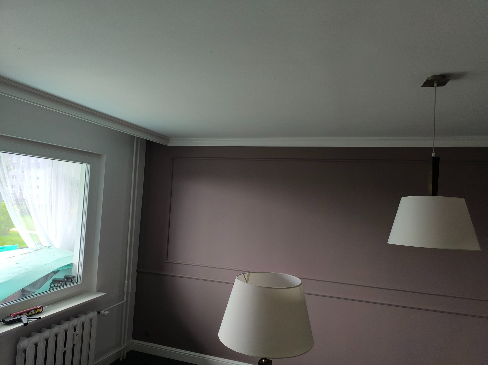 Ściana w odcieniach fioletu z dekoracyjnymi listwami, okno z zasłoną i zabezpieczeniem, lampy. Widoczne narzędzia na parapecie. Wykończenie wnętrza.