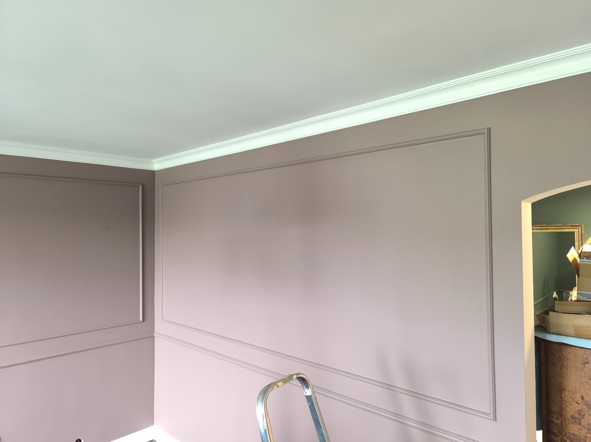 Ściana z dekoracyjnymi listwami pomalowana na liliowo w Szczecinie. Widoczny fragment drabiny i kartony w tle. Klasyczny wystrój wnętrza po renowacji.