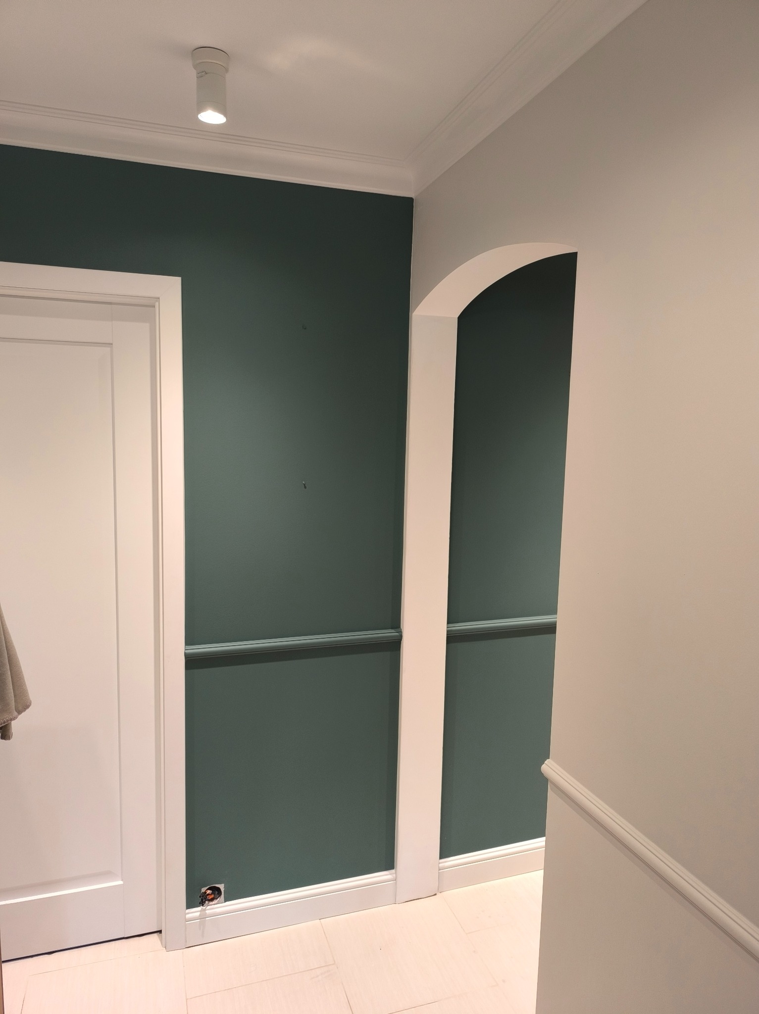 Korytarz po remoncie: ściana w kolorze butelkowej zieleni z białą sztukaterią, białe drzwi i łukowate przejście. Widoczna podłoga z jasnych płytek i punktowe oświetlenie.