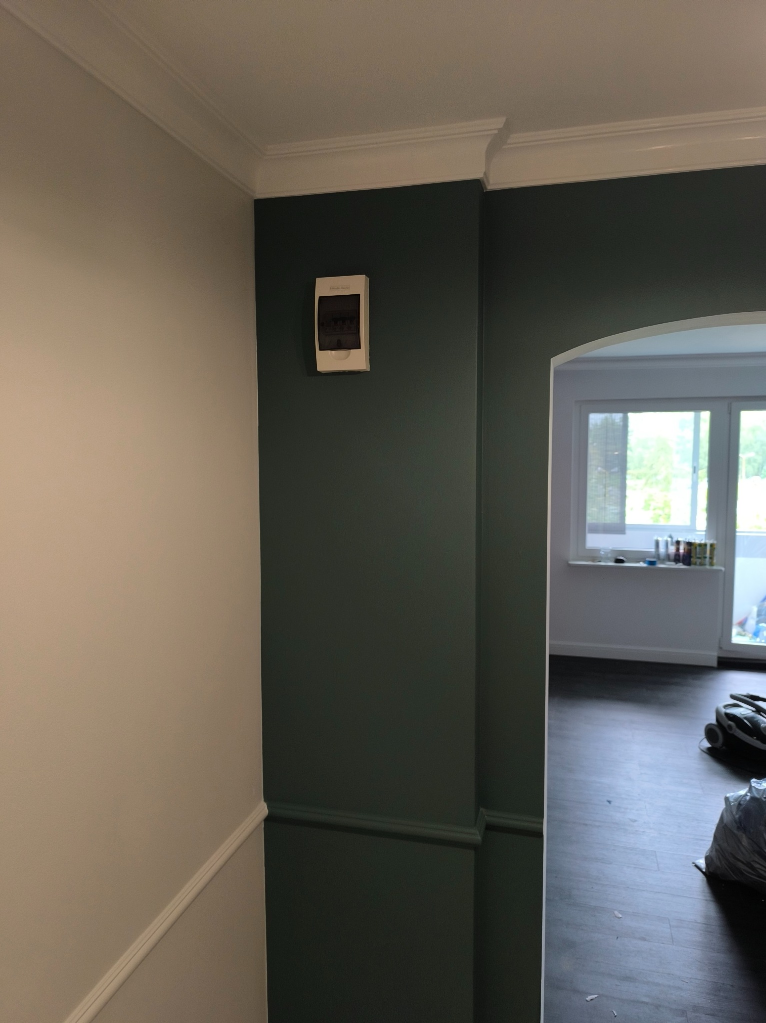 Ściana pomalowana na dwa kolory z dekoracyjną listwą i skrzynką elektryczną. Widok na pokój z oknem i podłogą z paneli. Częściowo widoczny odkurzacz.