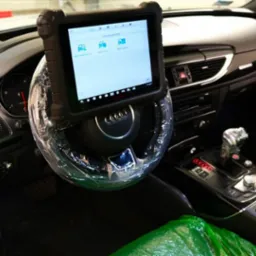 Wnętrze Audi z podłączonym tabletem diagnostycznym na kierownicy, osłoniętej folią. Widoczny panel środkowy i dźwignia zmiany biegów.