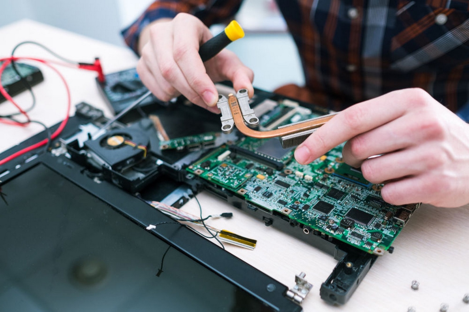 Naprawa laptopa: osoba w koszuli w kratę demontuje płytę główną, używając śrubokręta. Widoczne elementy: radiator, wentylator, kable. Jasne oświetlenie, precyzyjne ujęcie.