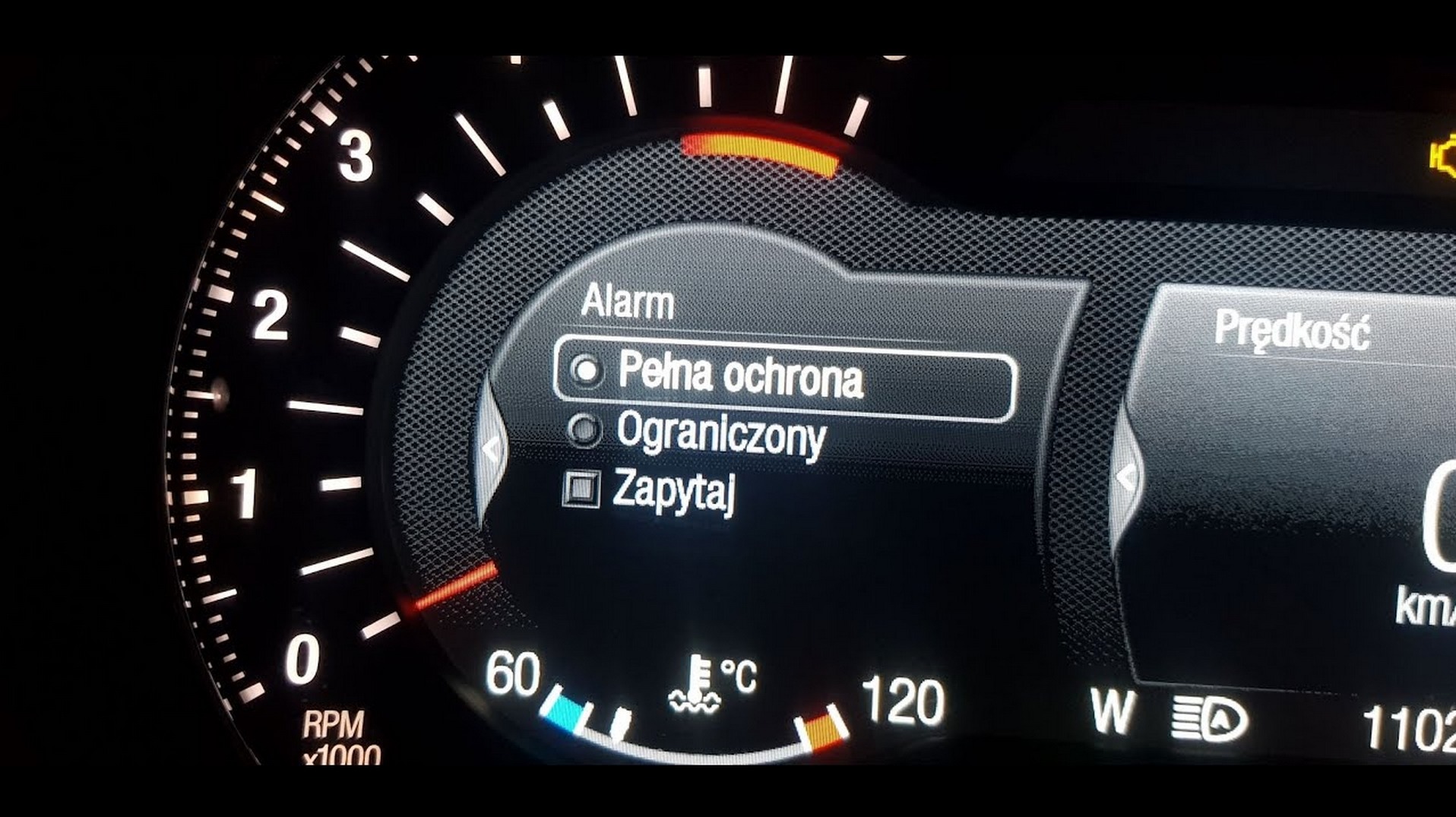 Deska rozdzielcza auta z menu alarmu: pełna ochrona, ograniczony, zapytaj. Widoczny obrotomierz, temperatura, prędkość i kontrolka silnika.