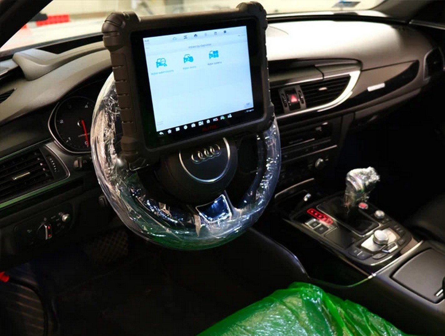 Wnętrze Audi z podłączonym tabletem diagnostycznym na kierownicy, osłoniętej folią. Widoczny panel środkowy i dźwignia zmiany biegów.