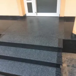 Granitowe schody wejściowe z dwoma kolumnami po bokach, prowadzące do białych drzwi. Powierzchnia błyszcząca, odbija światło.