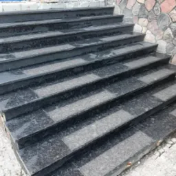 Granitowe schody zewnętrzne z ciemnego kamienia, wykończone z dbałością o detale, prowadzące do kamiennej ściany. Widoczny fragment kostki brukowej.