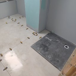 Rafkam - Układanie jasnych płytek podłogowych z użyciem poziomujących klipsów i czarnego brodzika w narożnej łazience z surowymi ścianami.