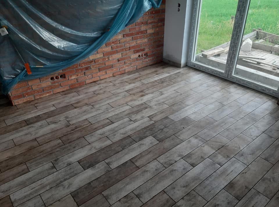 Ukończona podłoga z płytek imitujących drewno w nowym wnętrzu. Czerwona cegła na ścianie częściowo zakryta folią ochronną. Widok na zewnątrz przez okno.