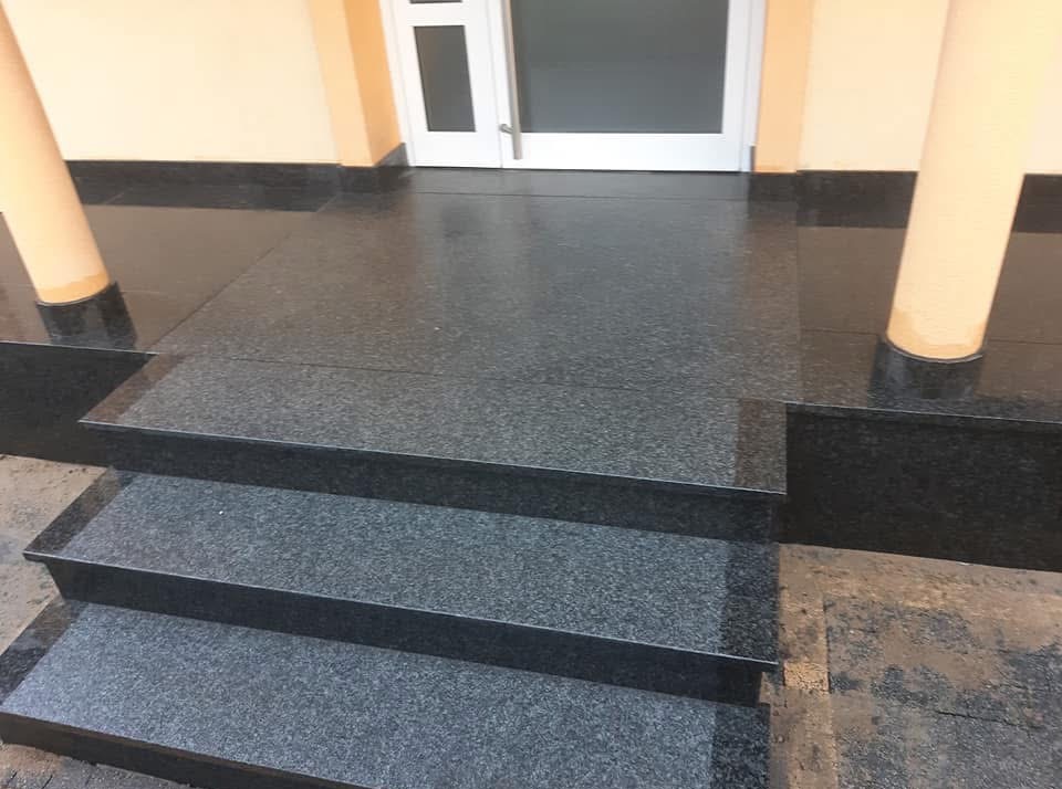 Granitowe schody wejściowe z dwoma kolumnami po bokach, prowadzące do białych drzwi. Powierzchnia błyszcząca, odbija światło.