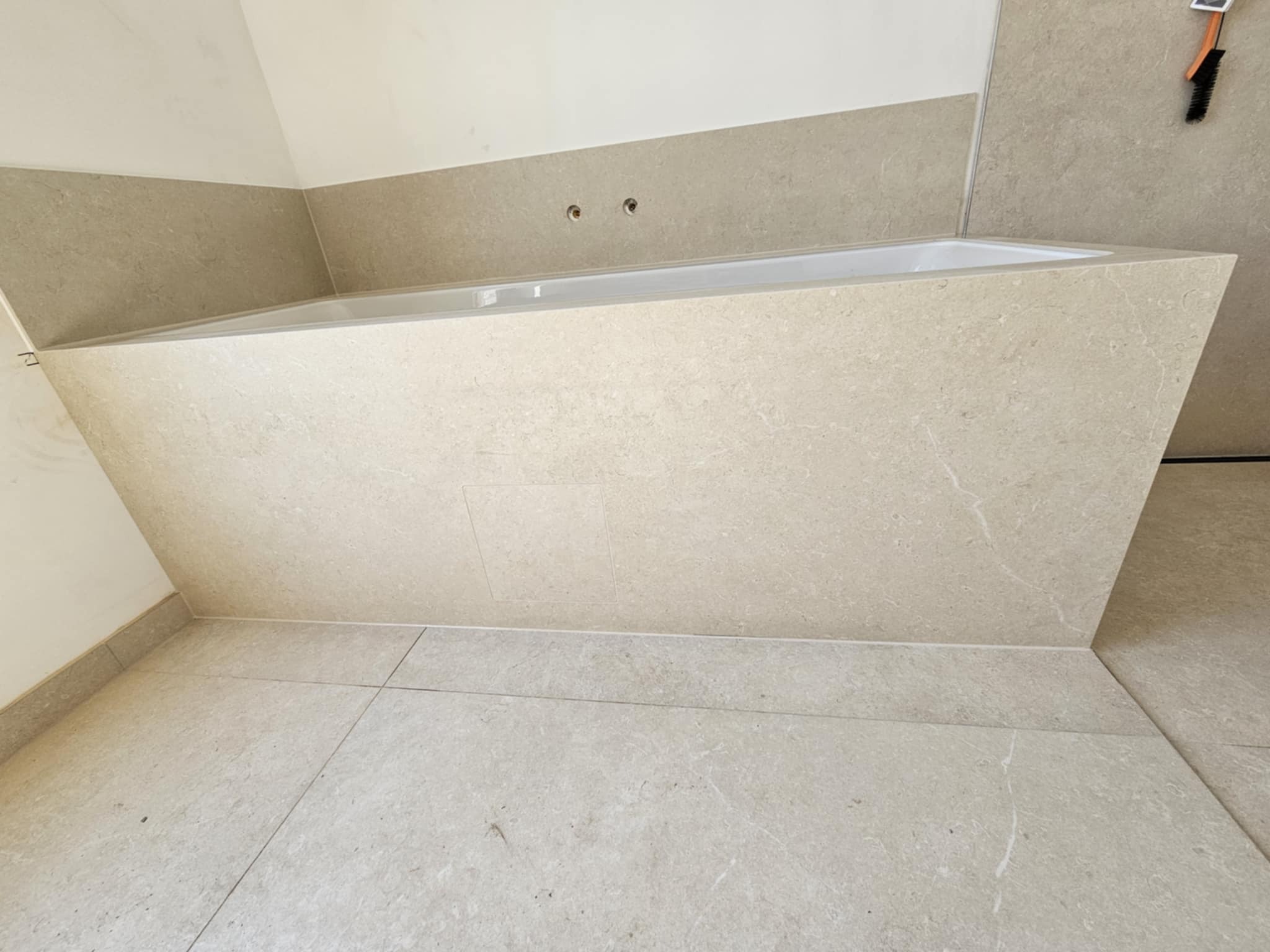 Wykończona beżowa wanna obłożona płytkami, z widocznymi fugami i fragmentem podłogi z tego samego materiału. Minimalistyczny design, surowe wykończenie.