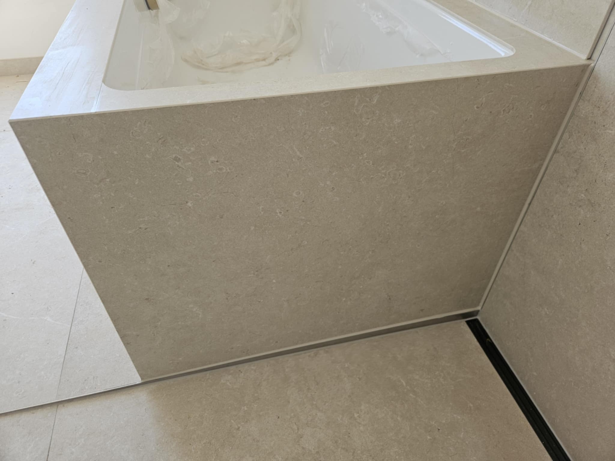 Obudowa wanny wykończona beżowymi płytkami imitującymi kamień, z widocznymi fugami i listwami wykończeniowymi. Minimalistyczny design, neutralna kolorystyka.