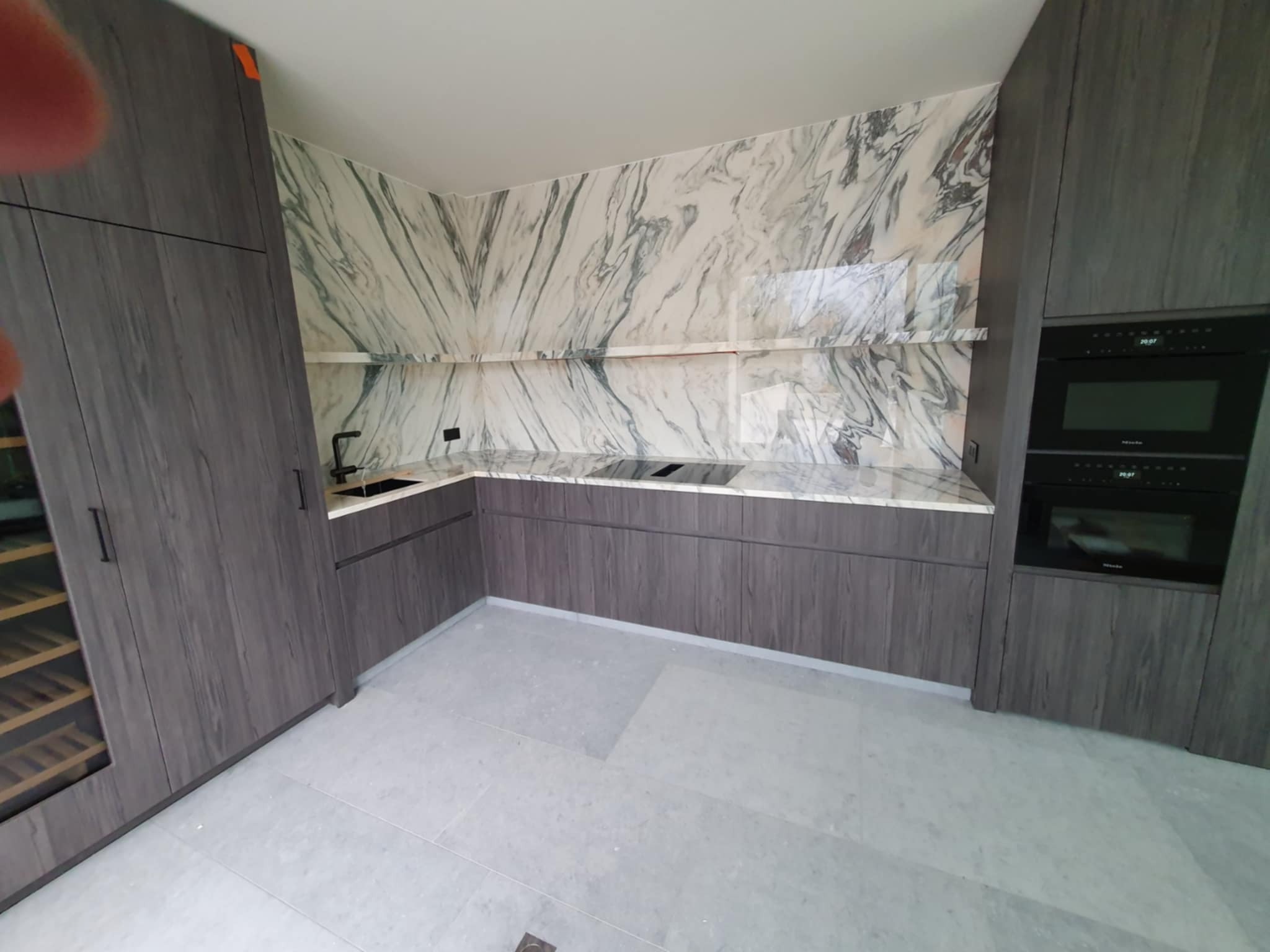 Nowoczesna kuchnia z marmurowym blatem i ścianą, szare szafki z drewnianą fakturą, wbudowane sprzęty AGD. Minimalistyczny design, jasne płytki podłogowe.