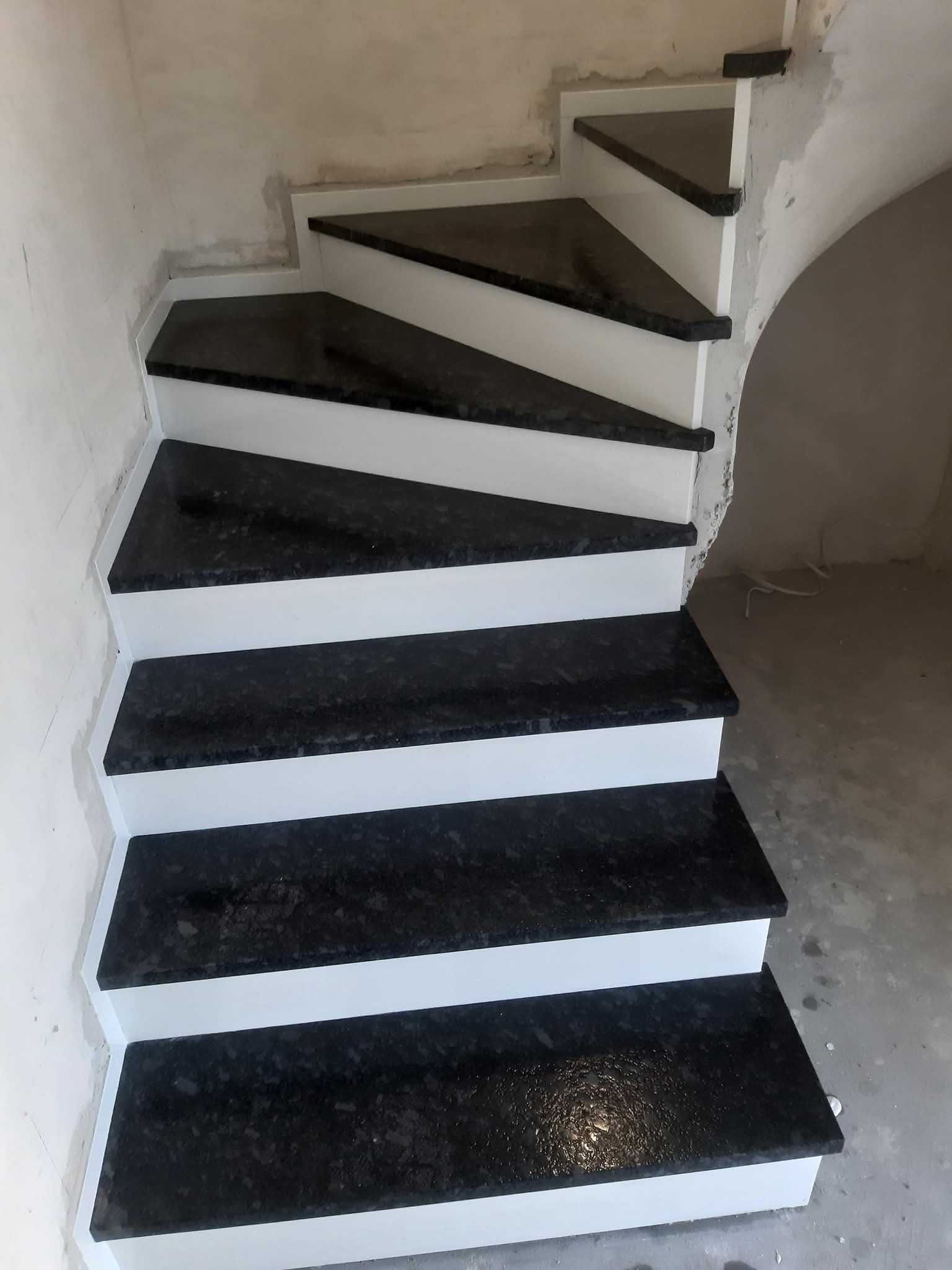 Wykończone schody z czarnego granitu i białych podstopnic, widok z dołu. Detal wykończenia w surowym wnętrzu, zarys łuku w tle.