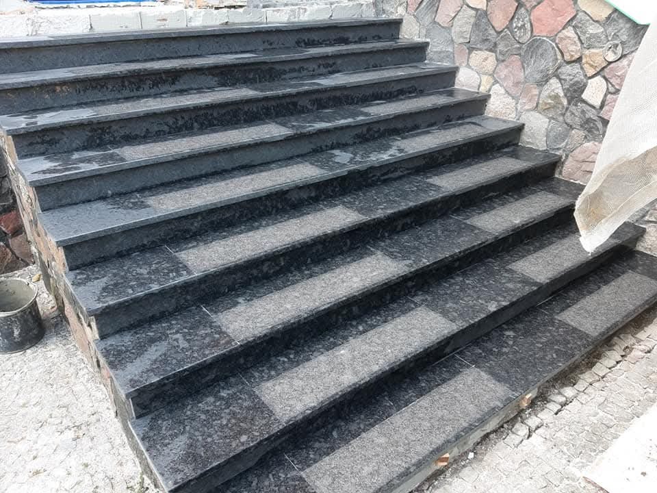 Granitowe schody zewnętrzne z ciemnego kamienia, wykończone z dbałością o detale, prowadzące do kamiennej ściany. Widoczny fragment kostki brukowej.