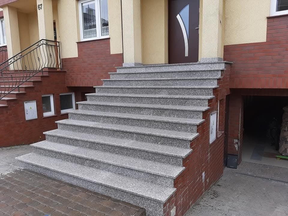 Granitowe schody wejściowe do budynku z numerem 67, wykończone czerwoną cegłą. Widoczny fragment balkonu z czarną, kutą balustradą i wejście do garażu.
