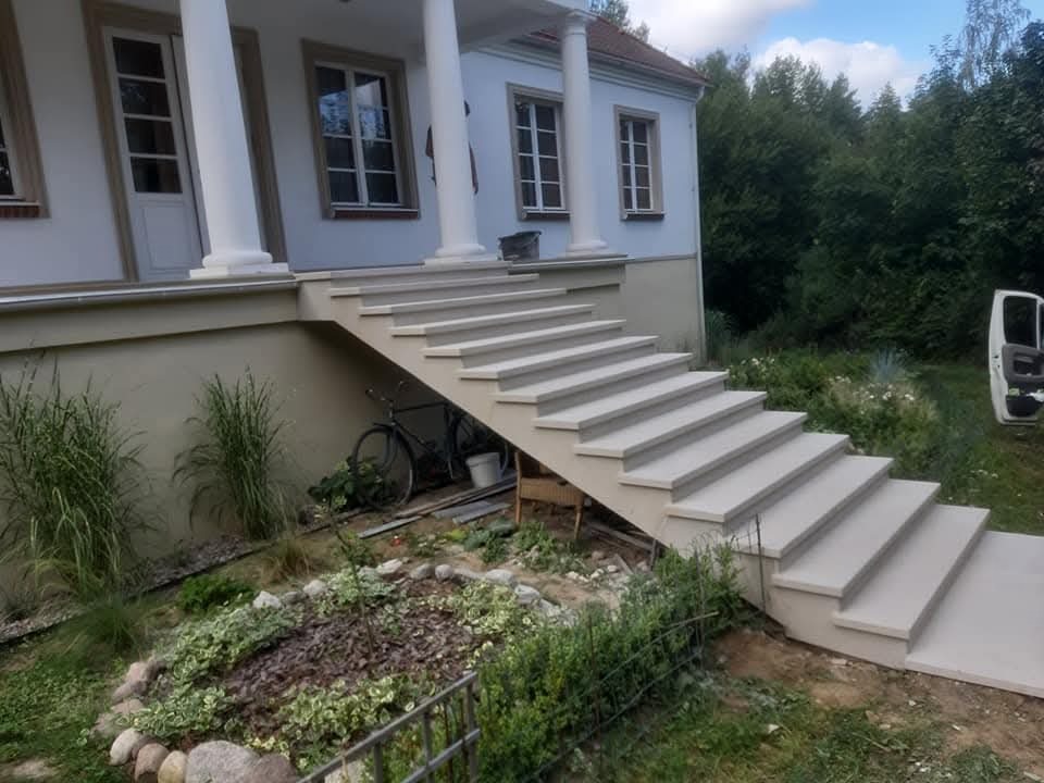 Wykończone schody zewnętrzne z jasnoszarej kostki prowadzące do domu z kolumnami, widok na ogród i częściowo otwarte drzwi samochodu dostawczego.