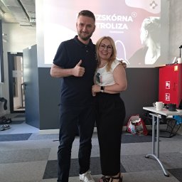 FizjoMed Gabinet Fizjoterapii i hirudoterapii mgr Gabriela Dąbrowiecka - Uśmiechnięty mężczyzna z uniesionym kciukiem i kobieta w okularach pozują na tle ekranu z napisem 'Przeszkórna Kontroliza Warszawa'.