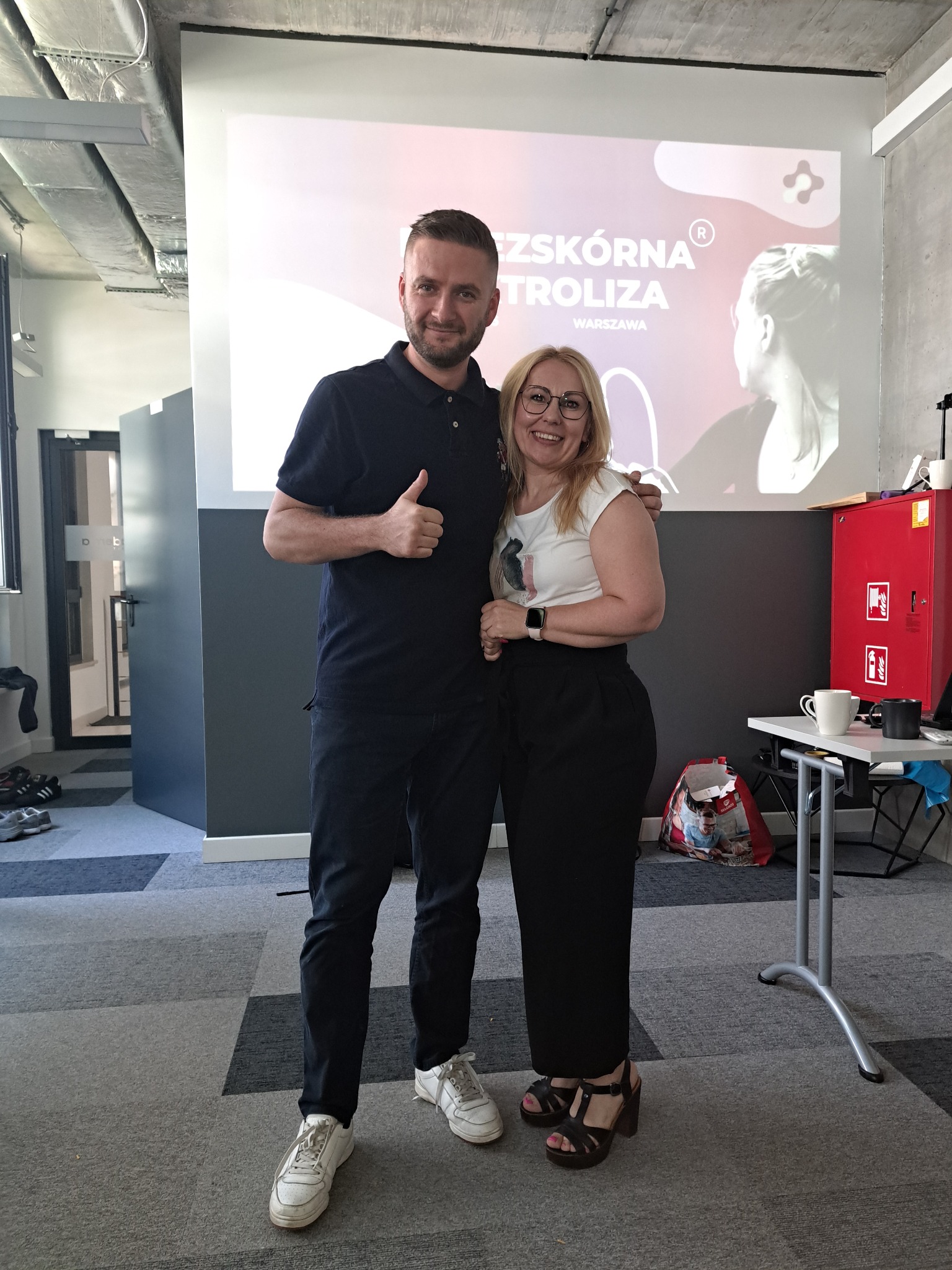 Uśmiechnięty mężczyzna z uniesionym kciukiem i kobieta w okularach pozują na tle ekranu z napisem 'Przeszkórna Kontroliza Warszawa'.