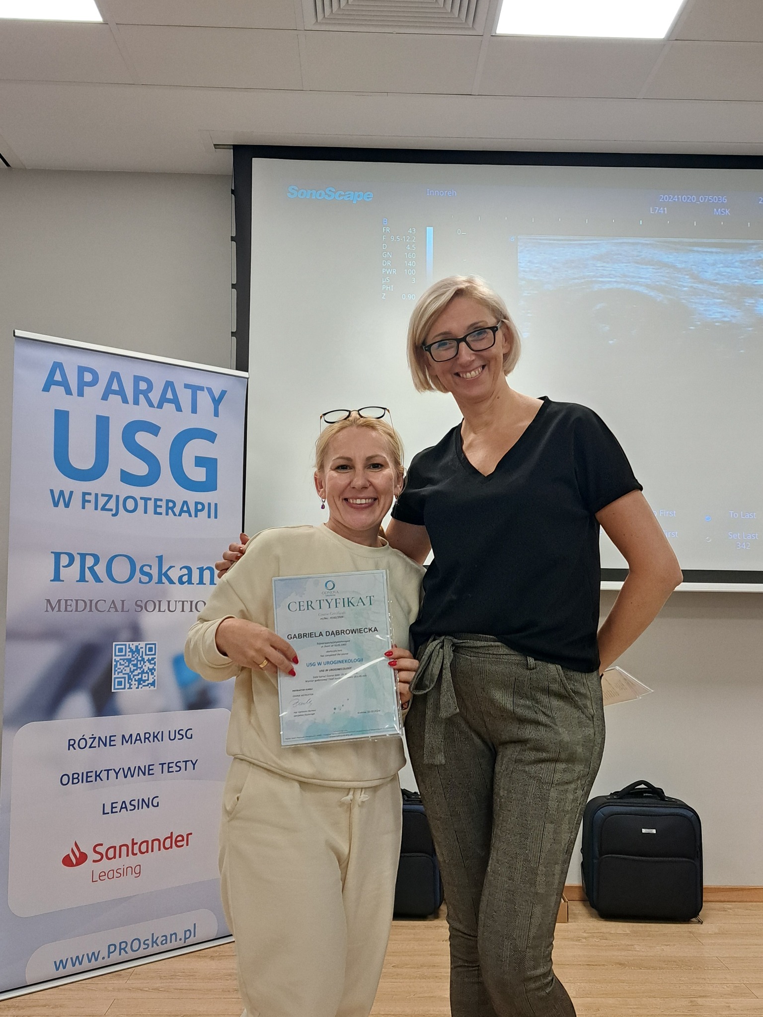 Dwie uśmiechnięte kobiety pozują z certyfikatem USG w uroginekologii, na tle ekranu z obrazem USG i banera z aparatami USG. Widoczny sprzęt medyczny.