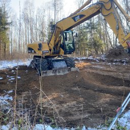 Mykyta Burlakov - Żółta koparka CAT przygotowuje teren pod budowę w lesie, widoczne ślady opon i resztki śniegu. Operator w kabinie, osoba z niwelatorem w tle.