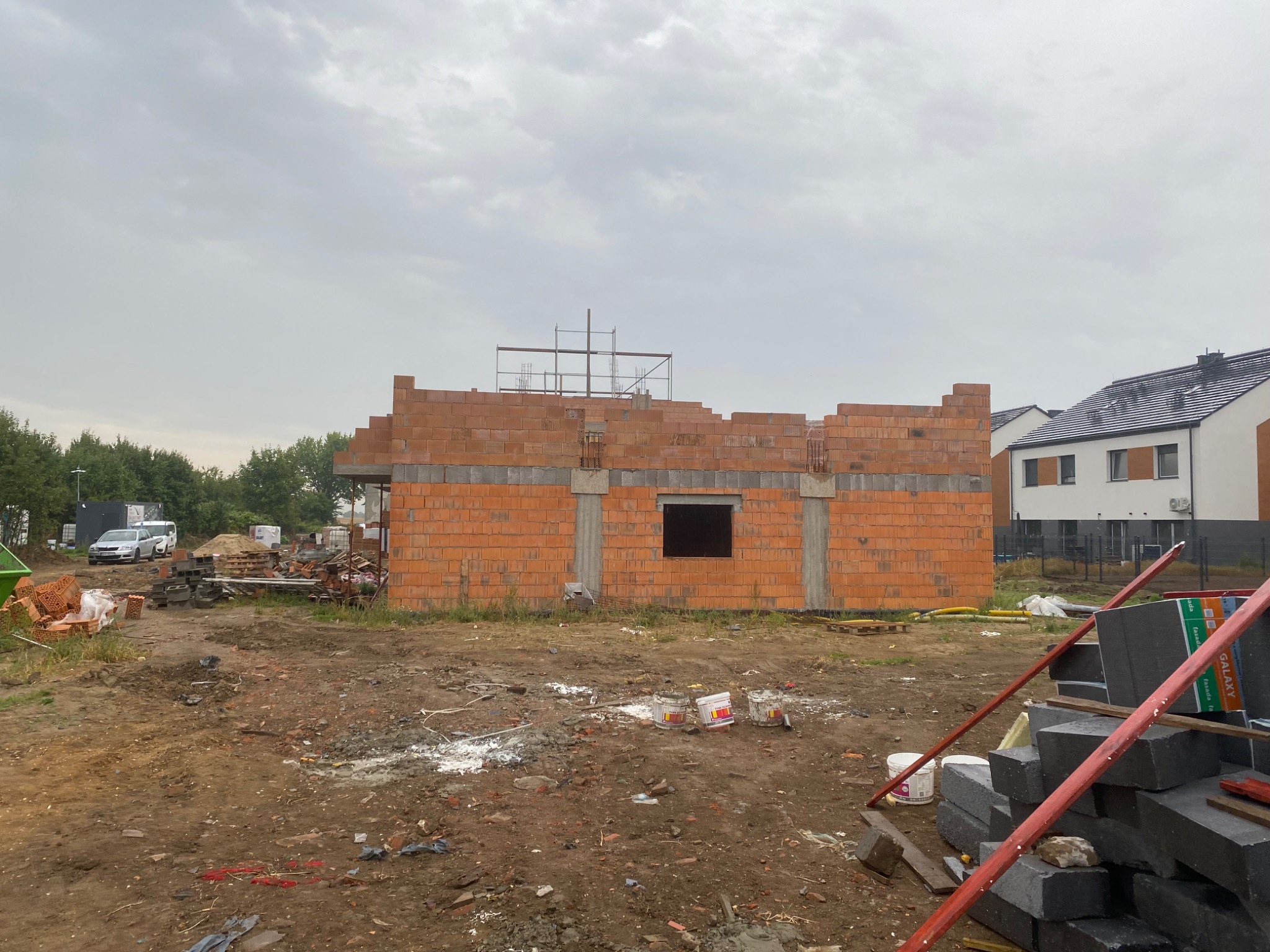 Budowa domu z czerwonej cegły w trakcie realizacji, widoczne rusztowania i materiały budowlane. Szary dzień, ziemiste podłoże.