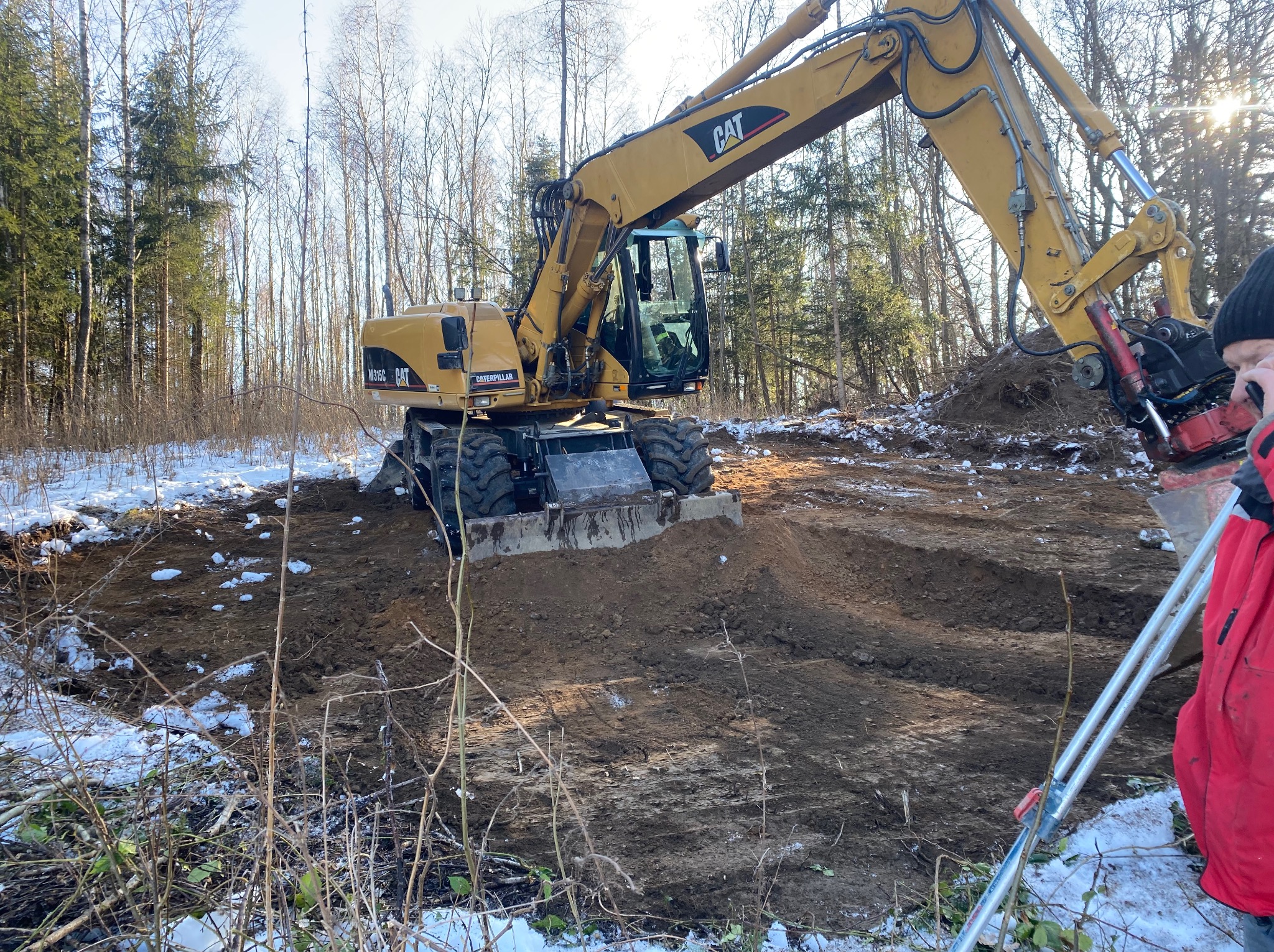 Żółta koparka CAT przygotowuje teren pod budowę w lesie, widoczne ślady opon i resztki śniegu. Operator w kabinie, osoba z niwelatorem w tle.