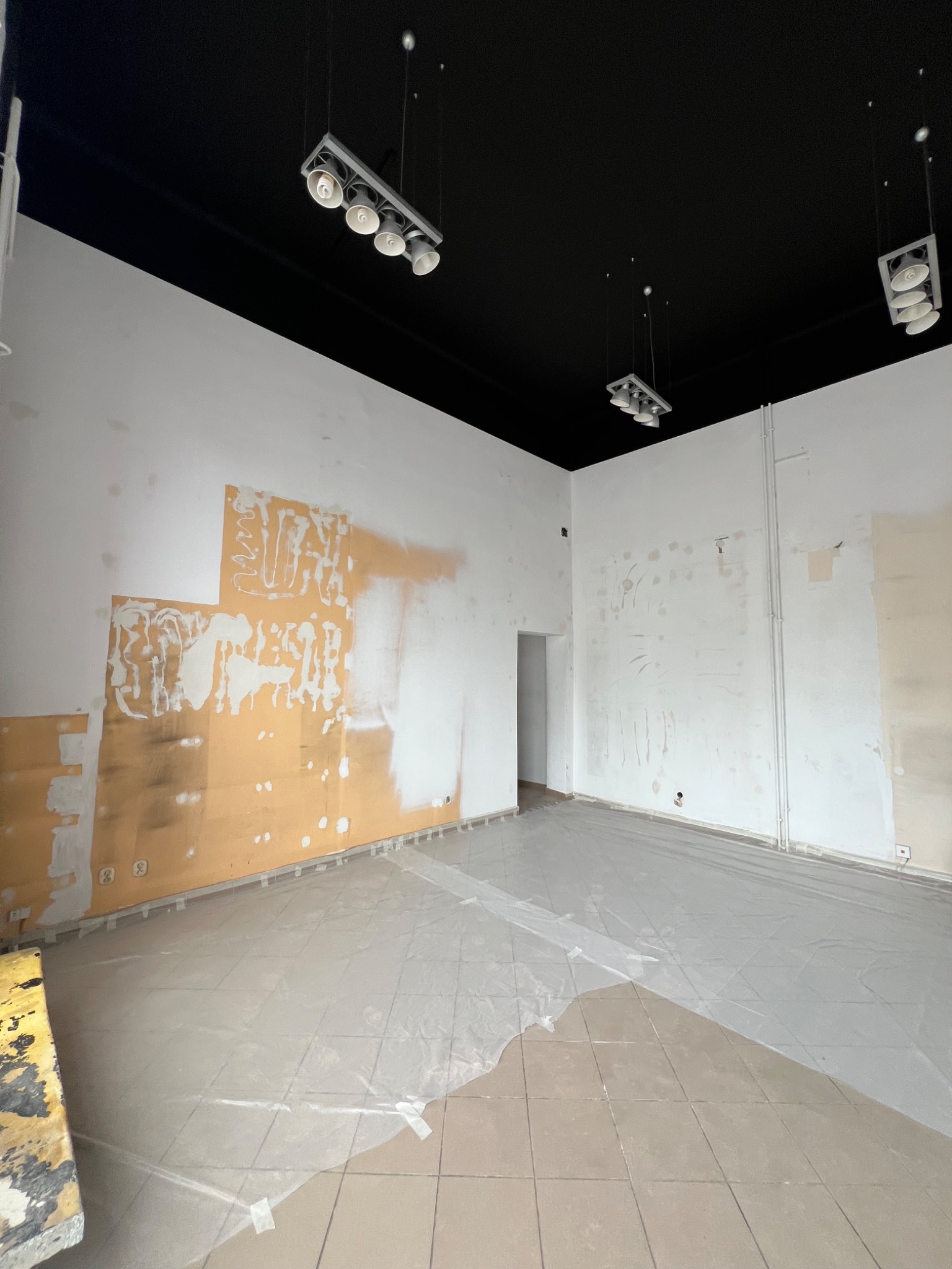 Wnętrze w trakcie remontu: przygotowana ściana z widocznymi śladami po szpachlowaniu, zabezpieczona podłoga folią, czarny sufit z oświetleniem.