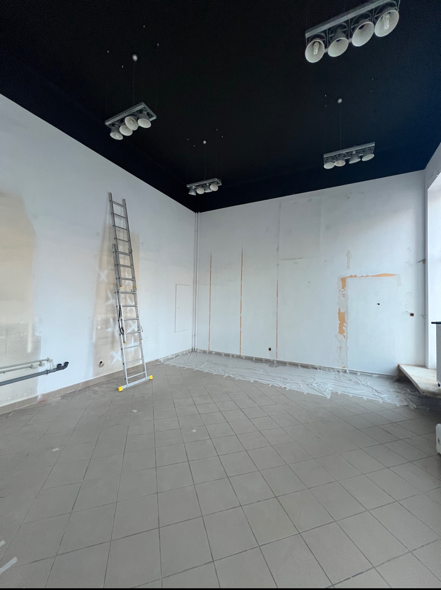 Przygotowane do malowania wnętrze biura: białe ściany, czarny sufit, drabina oparta o ścianę, podłoga zabezpieczona folią malarską, widoczne lampy sufitowe.