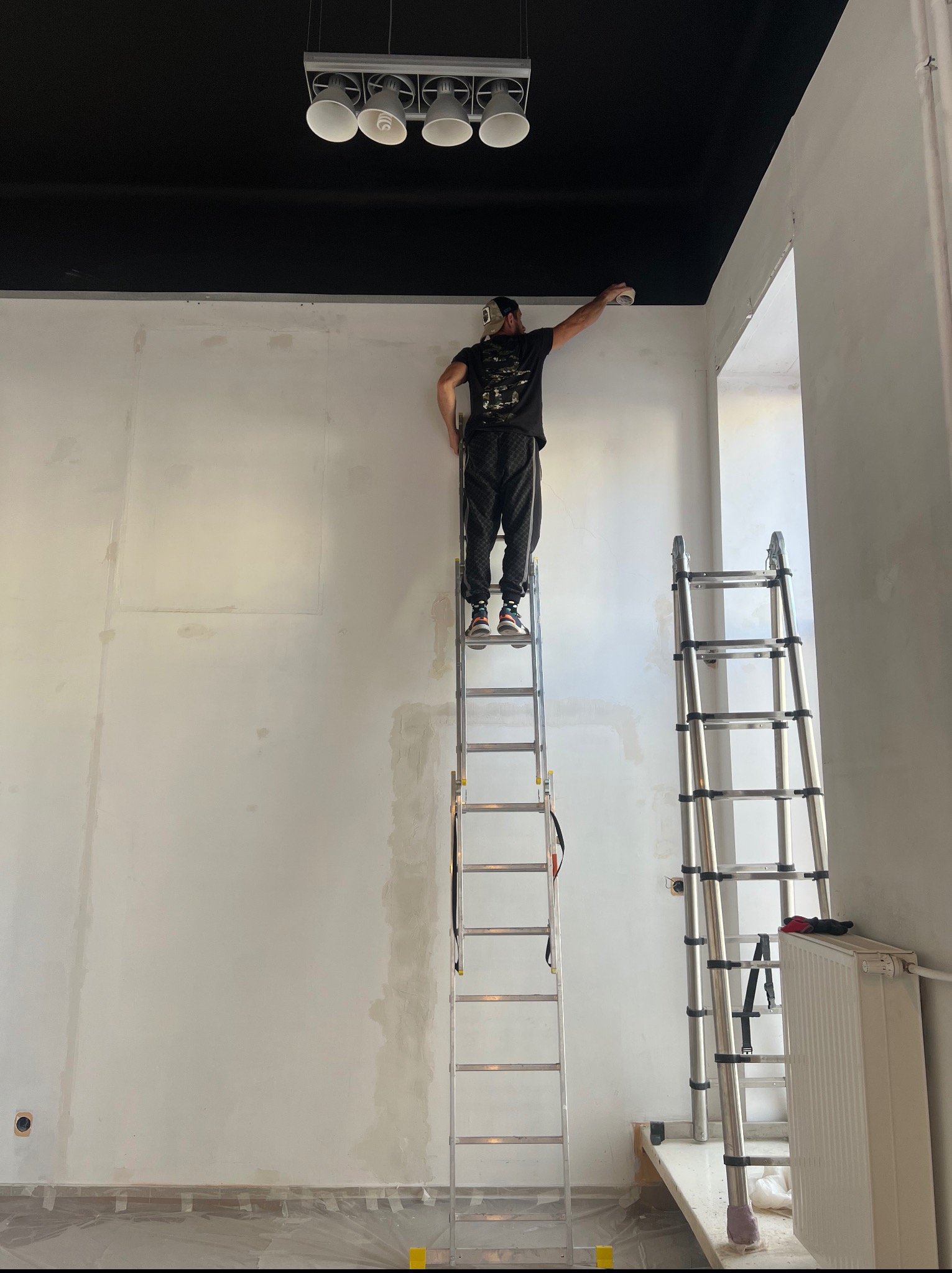 Mężczyzna na drabinie maluje sufit na czarno w jasnym pomieszczeniu. Widoczne dwie drabiny, zabezpieczona podłoga i częściowo pomalowana ściana.