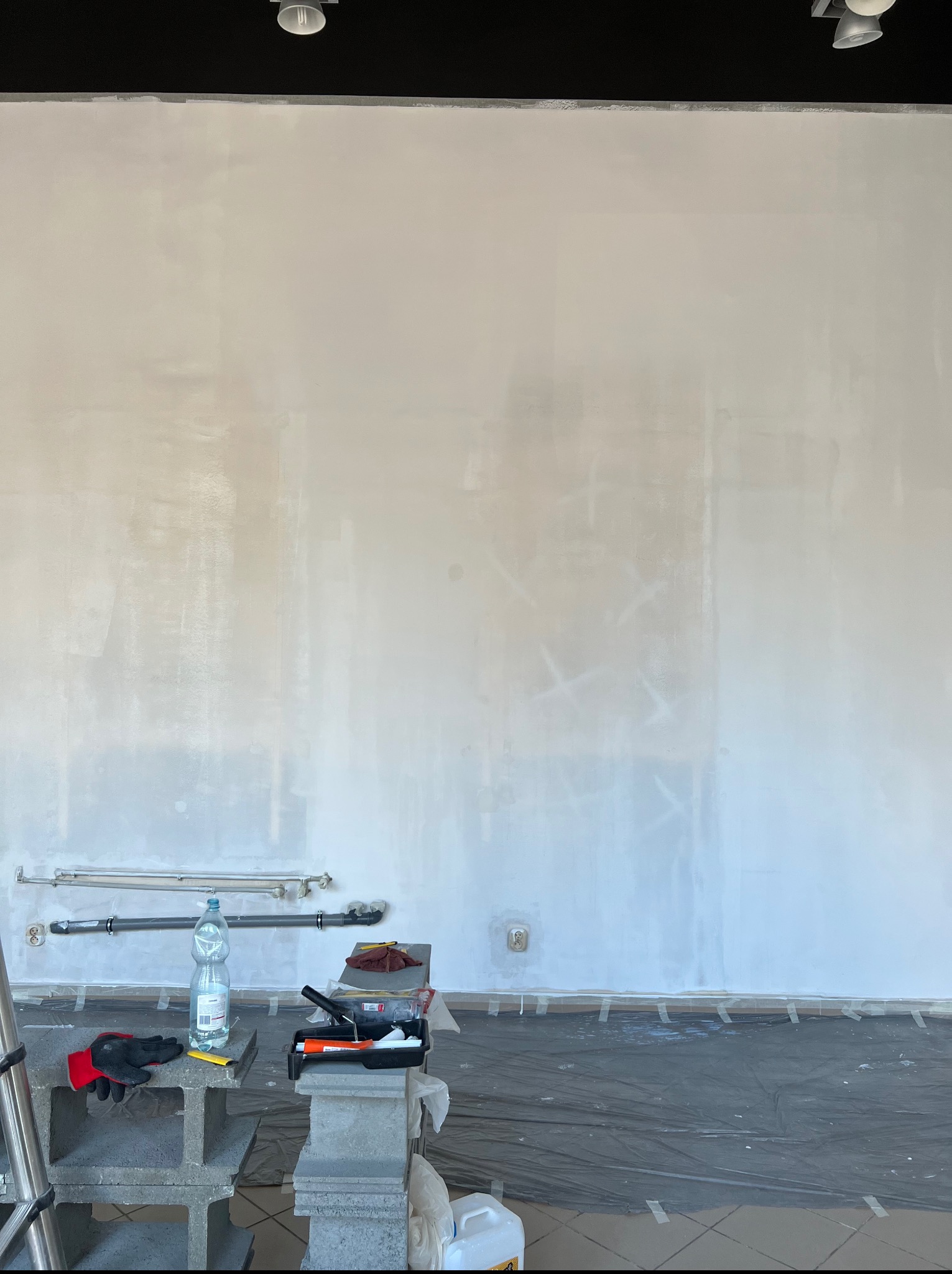 Ściana po gruntowaniu, przygotowana do malowania. Widoczne narzędzia malarskie, folia ochronna na podłodze i fragment drabiny.
