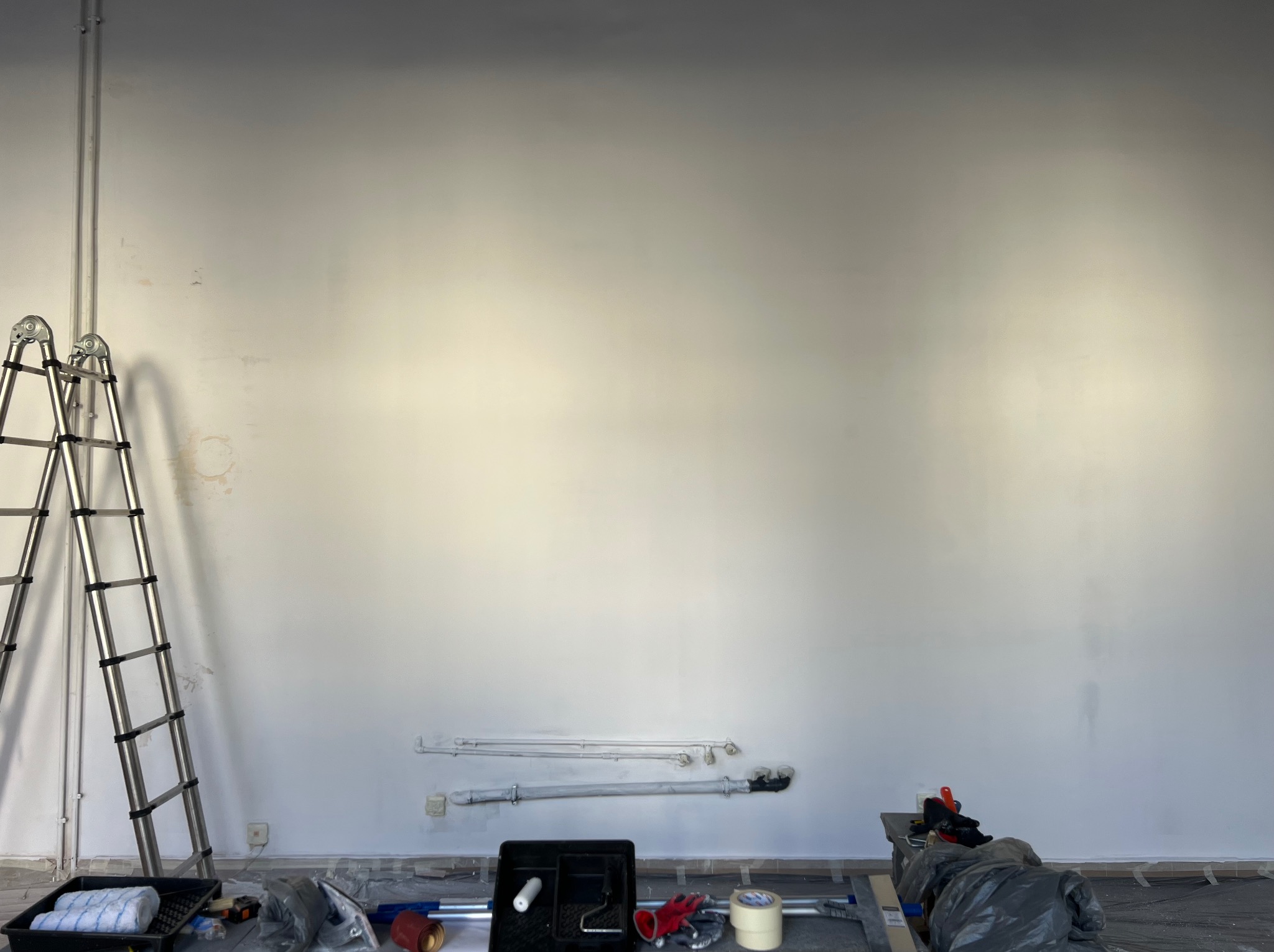 Ściana po malowaniu z widocznymi rurami i narzędziami malarskimi, drabina oparta o ścianę, podłoga zabezpieczona folią malarską.