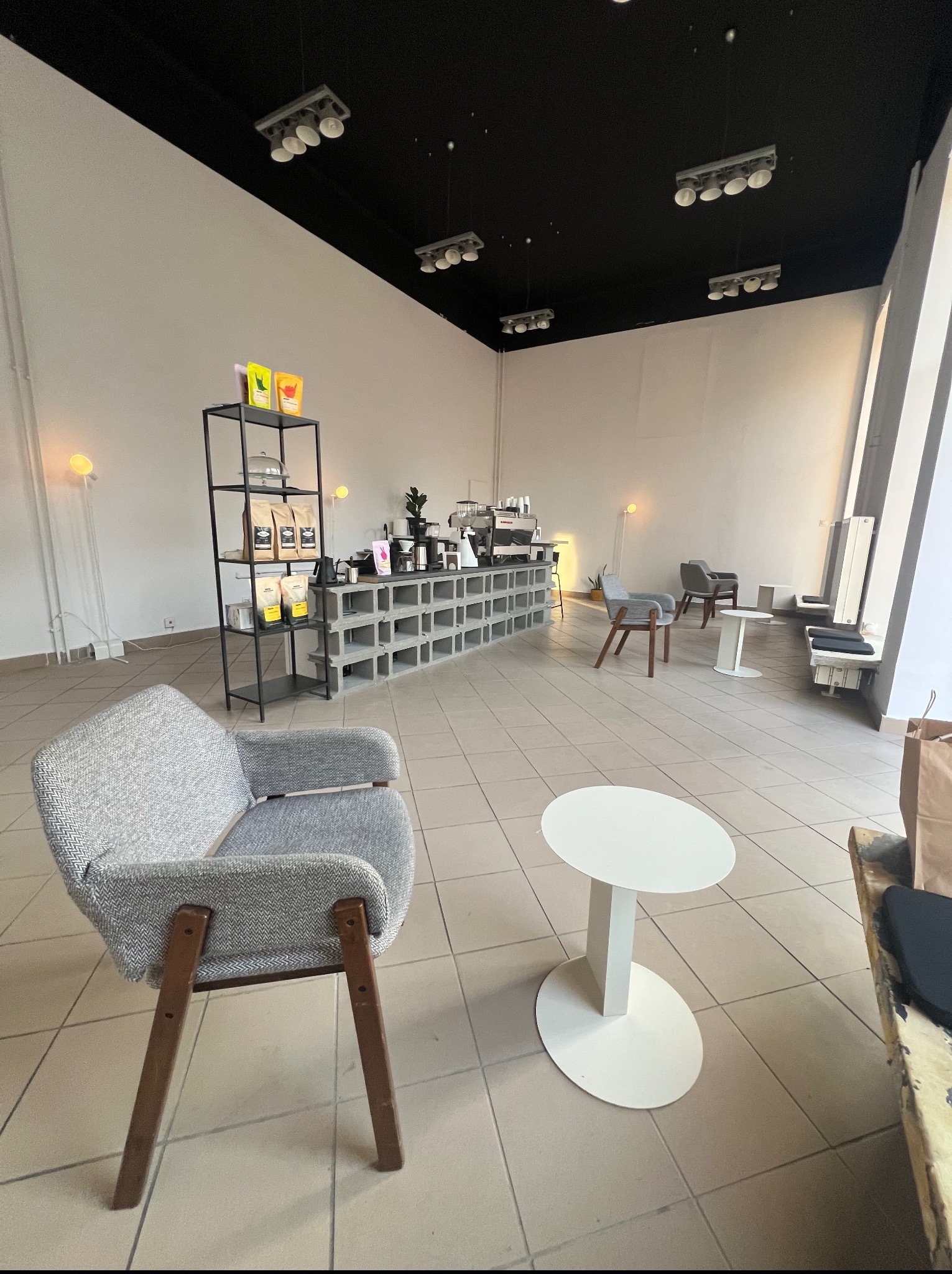 Nowoczesne wnętrze kawiarni z szarymi fotelami, białymi stolikami i betonowym barem. Czarno-biała kolorystyka, minimalistyczny design, światło dzienne z okna.