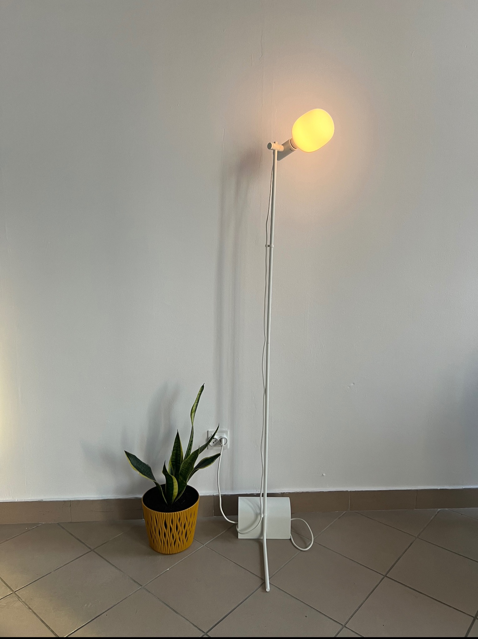 Minimalistyczna biała lampa podłogowa z żółtą żarówką i doniczką z sansewierią na tle gładkiej, jasnej ściany. Widoczny fragment gniazdka elektrycznego i brązowe płytki podłogowe.