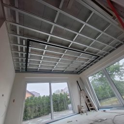 Oskar Czernecki - Sufit podwieszany z metalową konstrukcją w jasnym wnętrzu z dużymi oknami, drabina oparta o ścianę, widoczne przewody elektryczne.