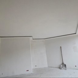 Oskar Czernecki - Wnętrze w trakcie wykończenia: ściany po szpachlowaniu, widoczne listwy przysufitowe, szpachla na podłodze, okno z widokiem na zieleń i cegły. Łopata oparta o ścianę.