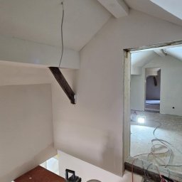 Oskar Czernecki - Wnętrze poddasza po szpachlowaniu i malowaniu na biało. Widoczne rusztowanie, narzędzia i lampa oświetlająca pomieszczenie. Elementy drewniane zachowane.