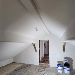 Oskar Czernecki - Poddasze po szpachlowaniu i malowaniu na biało, widoczne puszki z farbą, lampa robocza i kable na podłodze. Charakterystyczne skosy i belki stropowe.