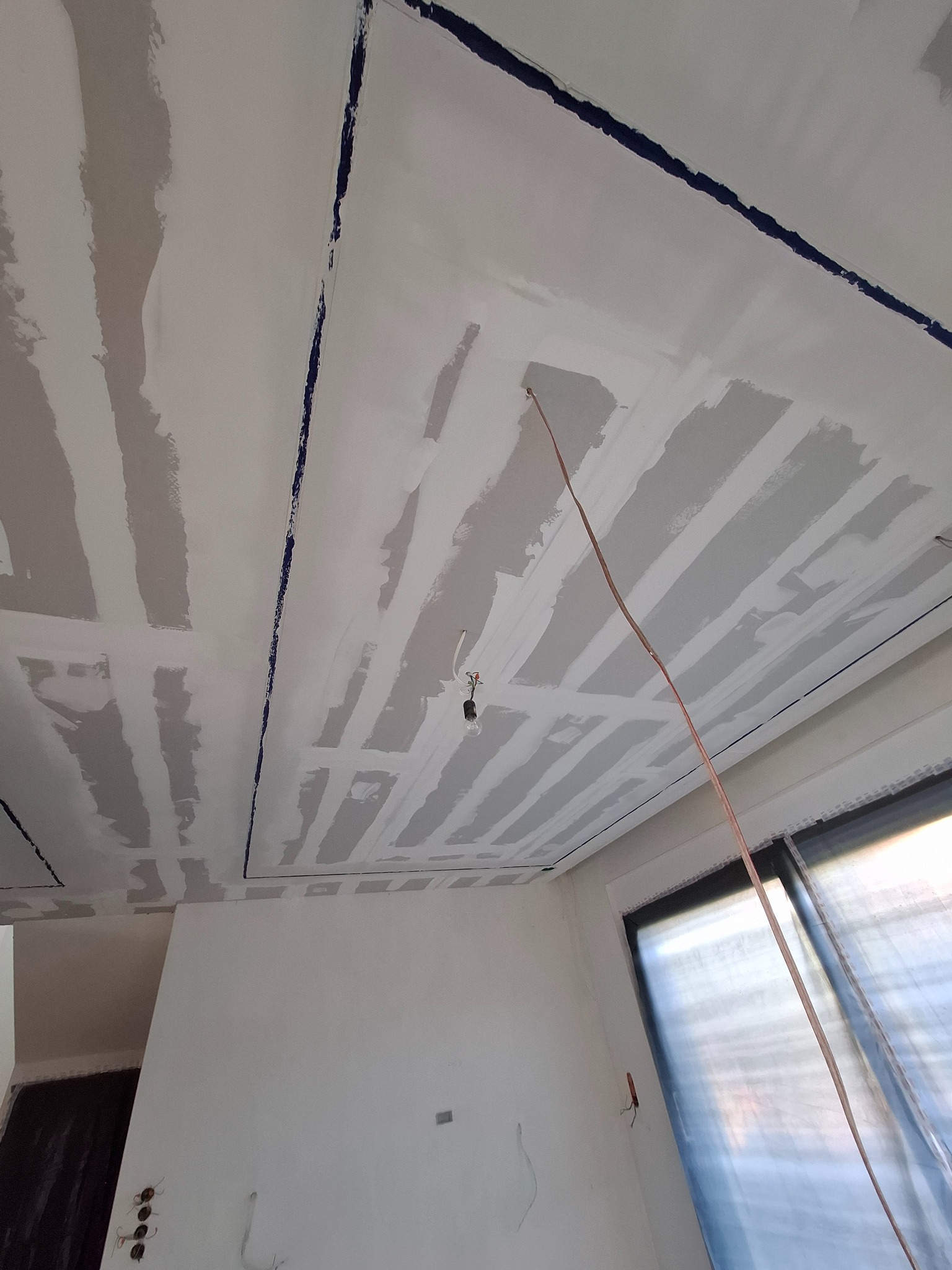 Sufit z płyt gipsowo-kartonowych w trakcie szpachlowania, widoczne łączenia i kable. Przy oknie folia ochronna. Ujęcie z dołu, sufit pod kątem.