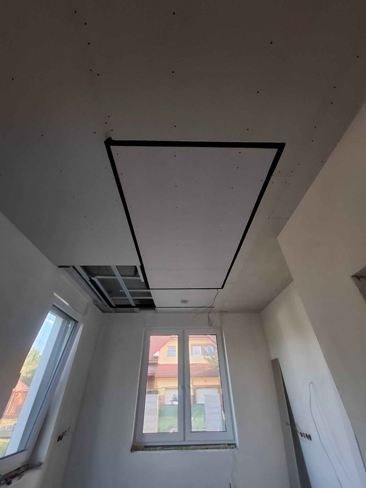 Częściowo zamontowany sufit z płyty gipsowej w jasnym pokoju z oknami. Widoczne łączenia płyt i metalowa konstrukcja. Etap prac wykończeniowych.