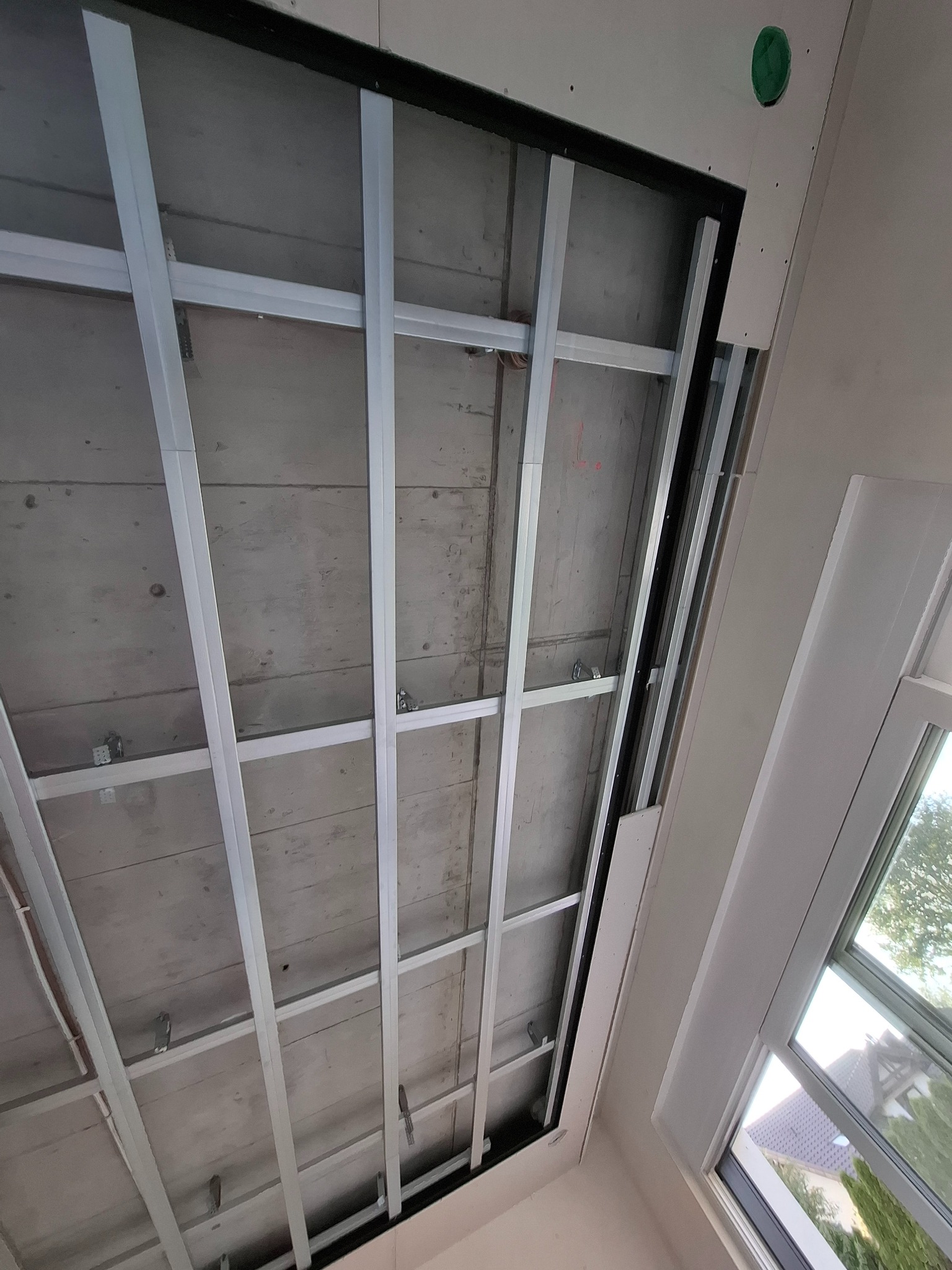 Montaż stelażu sufitu podwieszanego z płyt karton gips, widoczne profile metalowe i fragment okna dachowego. Etap prac przed montażem płyt.