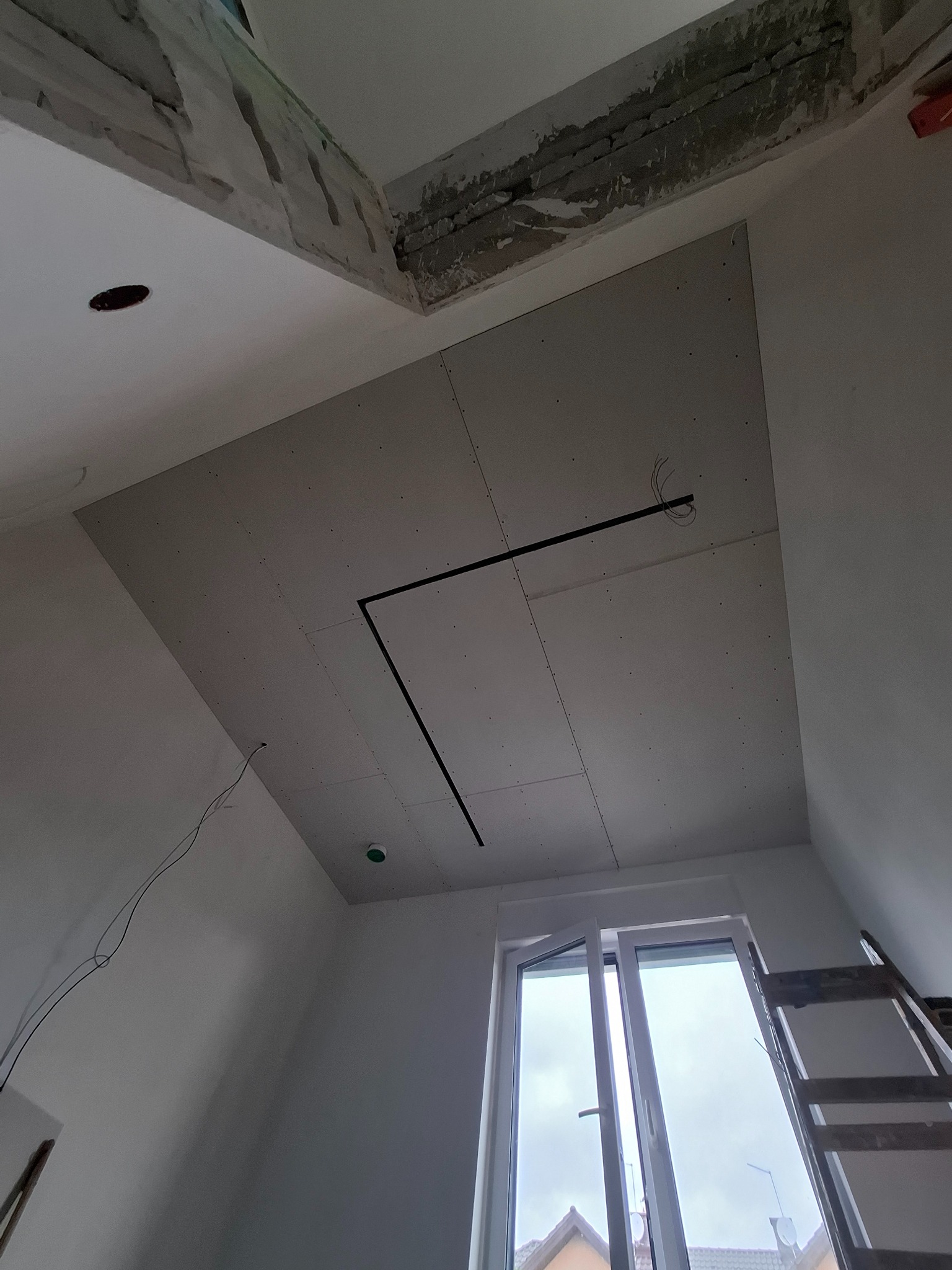 Montaż płyt gipsowo-kartonowych na suficie poddasza, widoczne profile, okno dachowe i drabina. Częściowo wykończone. Surowy stan.